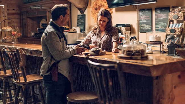 Johnathon Schaech and Sarah Lancaster engaged in conversation while standing at a cafe counter. Johnathon leans forward with his hands on the counter, while Sarah looks towards him with a smile. The cafe has a rustic decor with wooden panels and a chalkboard menu in the background.