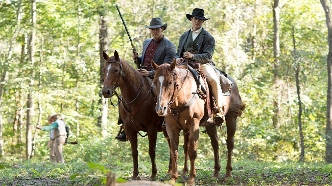 Neal McDonough and Steven R. McQueen on horseback in a rugged, wooded landscape with a green forest in the background