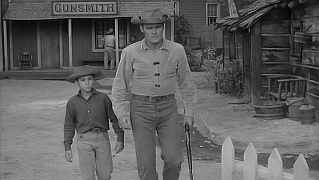 Chuck Connors and Johnny Crawford walking away from a Gunsmith shop down a road in town