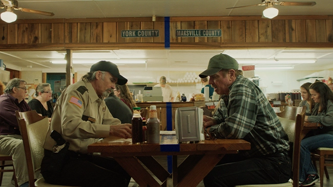 Tom Wopat and Jeff Fahey sitting across from each other at a table in a crowded local diner having a conversation.