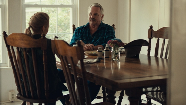 Tom Wopat sitting at a dining room table in a well-lit room, engaged in conversation with a woman.