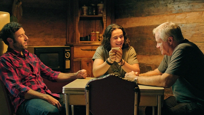David Keith, Coby Ryan McLaughlin, and Keean Johnson enjoying a game of cards in a cozy rustic cabin setting.