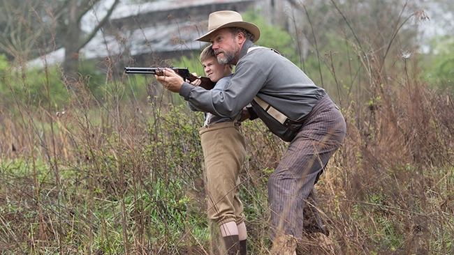 Adam Baldwin teaching Jet Jurgensmeyer how to shoot a firearm in a scenic outdoor field.