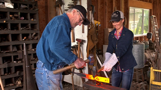 Jill Wagner showcasing her welding skills as she works on a metal object alongside a craftsman in a workshop.