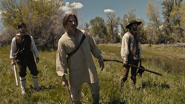 Three men dressed in period clothing, standing in a field with a stream in the background. The men are posed in a way that suggests they may be portraying historical figures from the 1800s.