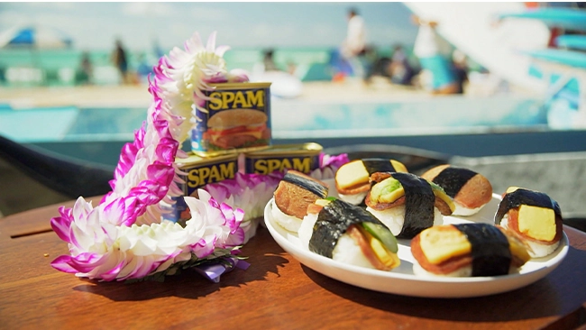 Stack of sushi and spam musubi with Hawaiian leis with a tropical background.