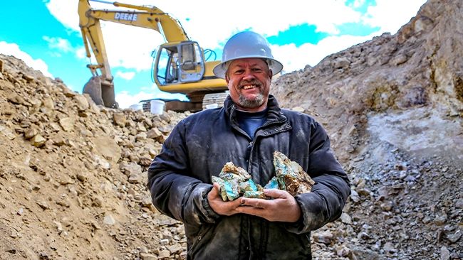 A scene from "Turquoise Fever," showcasing a cast member holding a handful of turquoise at a dig site. Heavy machinery and hard hats are used in the process of digging up these beautiful stones.